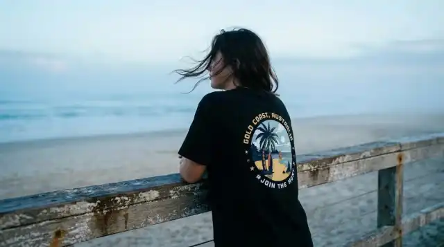 A woman wearing a premium 200gsm black cotton Sunny Escape souvenir t-shirt, featuring a circular Gold Coast Australia palm tree logo, looking out over a beach on the Gold Coast Australia at sunset.
